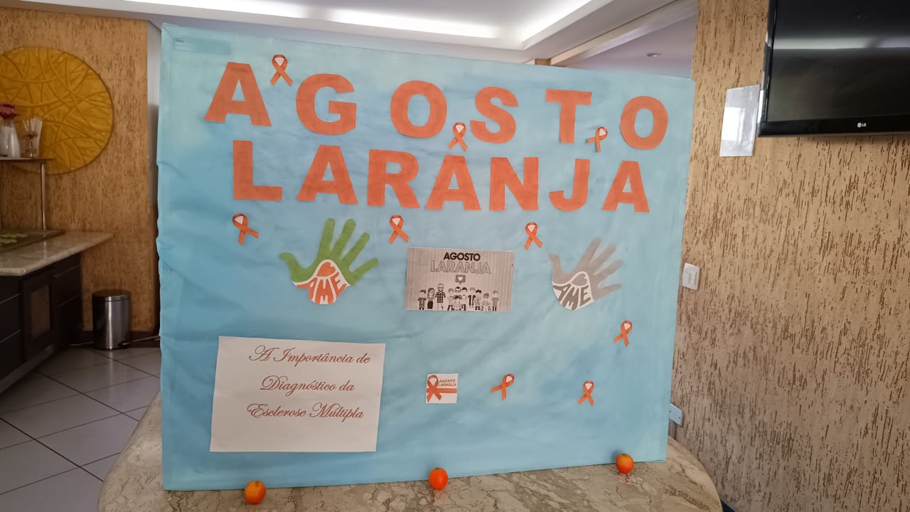 Na imagem, um cartaz azul com letras laranjas que escrevem: "Agosto Laranja". Mais abaixo, colada no cartaz, uma folha branca escrito: "A importância do diagnóstico da esclerose múltipla".